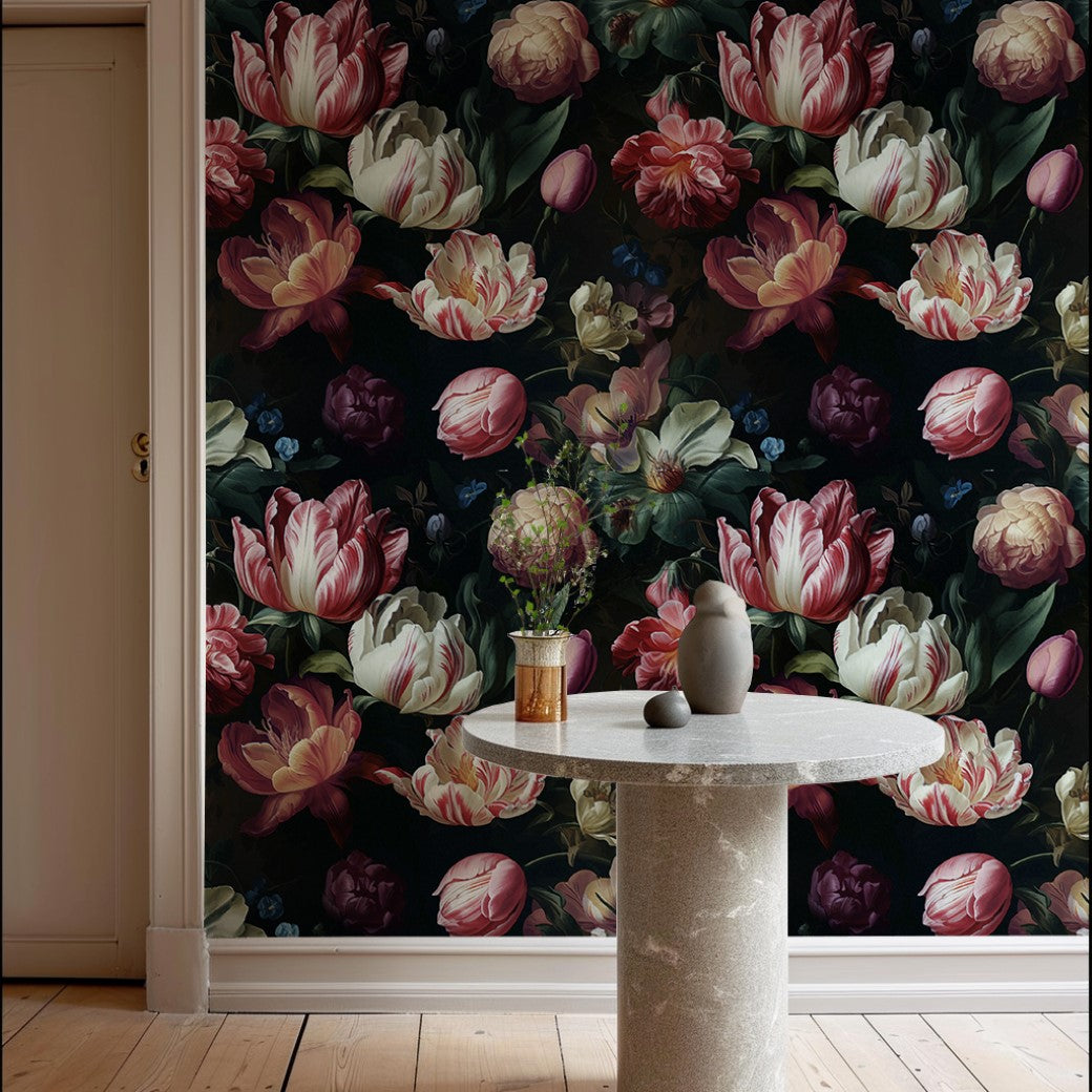 A round gray table sits before Decor2Go's Dark Elegance Floral Wallpaper Mural, featuring dark green leaves and vibrant flowers. On the table are a small vase with blooms and two sculpted figures. The light wood floor contrasts beautifully with the luxurious dark background.