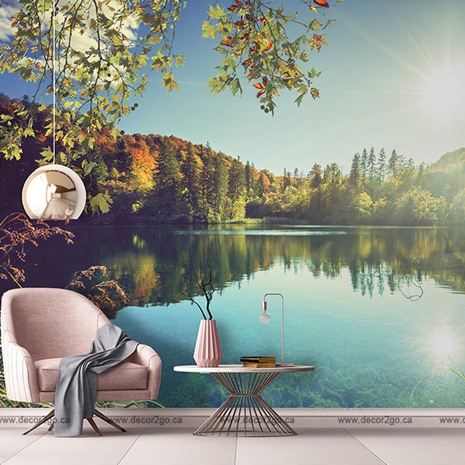 A serene lakeside scene with a pink armchair and small table, framed by lush trees and a clear blue sky, reflecting sunlight in a tranquil lake. A hanging lamp and a cozy blanket add comfort with the Day on The Lake Wallpaper Mural from Decor2Go Wallpaper Mural.