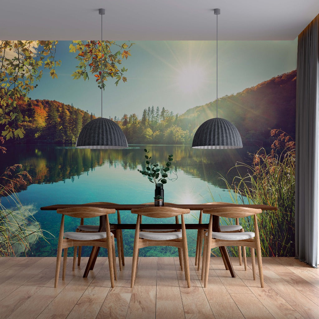 A modern dining area with wooden table and chairs, facing a large window with a vibrant Decor2Go Wallpaper Mural of a serene lake, surrounded by forest and reflecting the sunset.