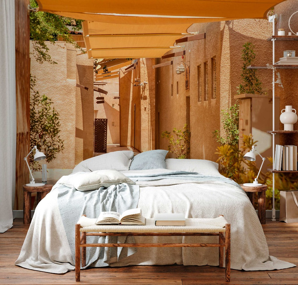 Bedroom - An outdoor bedroom setup in a narrow alley, featuring a large bed with blue and white bedding, wooden floor, and beige fabric shades above. A small bench with an open book is at the foot of Decor2Go Wallpaper Mural.