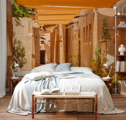 Bedroom - An outdoor bedroom setup in a narrow alley, featuring a large bed with blue and white bedding, wooden floor, and beige fabric shades above. A small bench with an open book is at the foot of Decor2Go Wallpaper Mural.