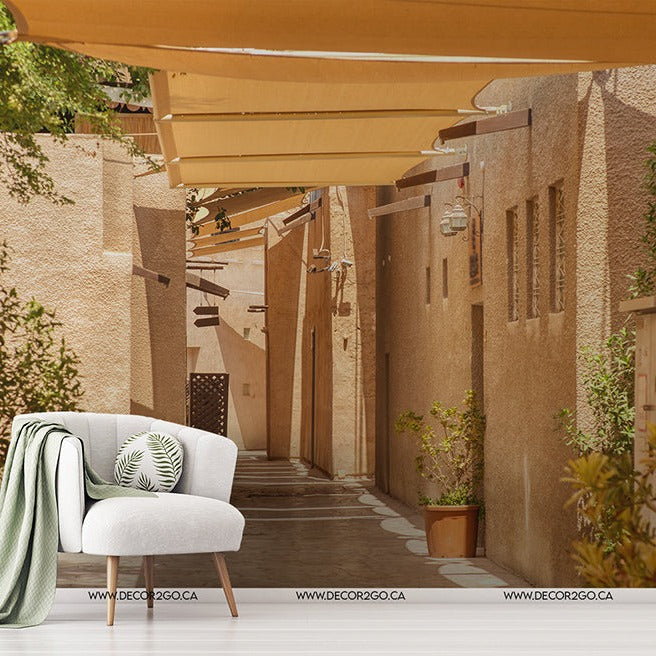 An inviting modern outdoor seating area with a white armchair and decorative cushions, nestled in a narrow alley with sandy walls and wooden beams, highlighted by potted plants and a sunlit ambiance, perfectly captured Decor2Go Wallpaper Mural Desert Alley.