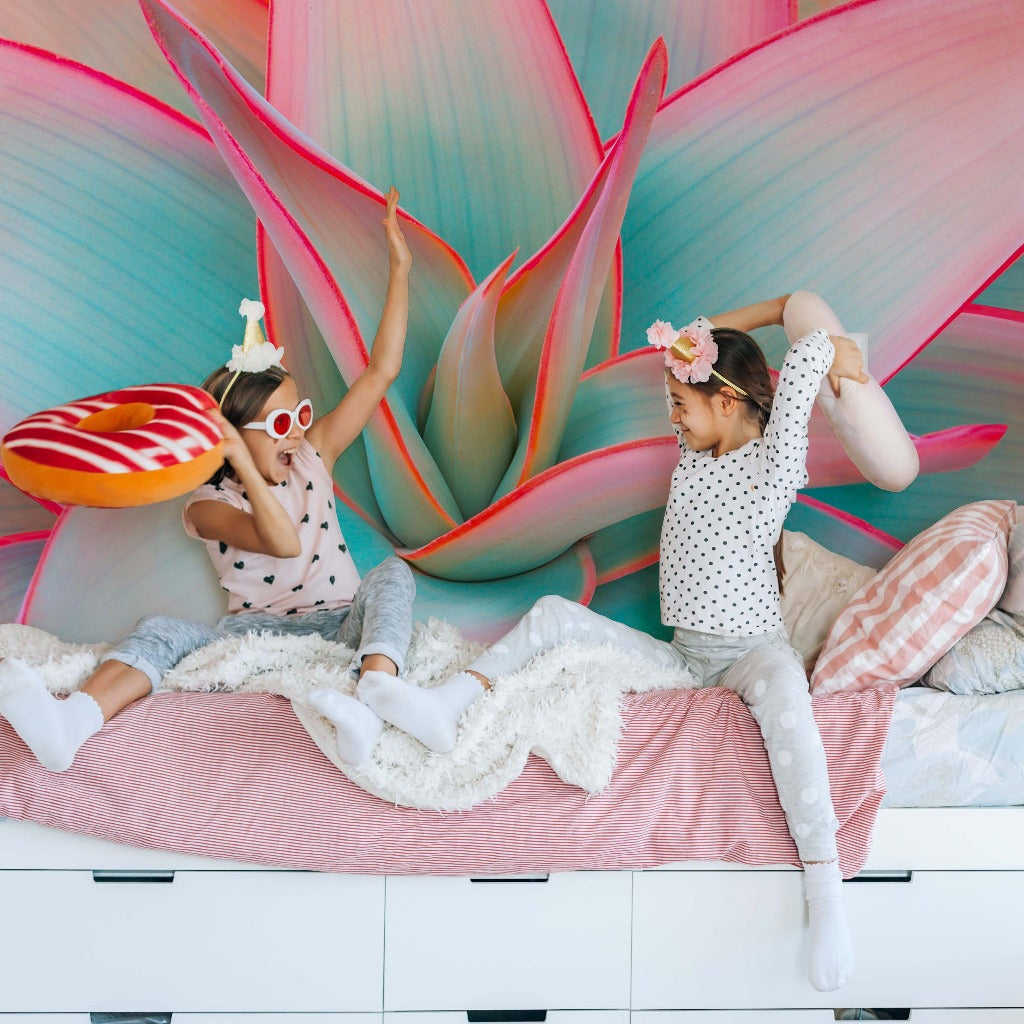 Two children having a pillow fight on a bed, with a vibrant Desert Oasis wallpaper mural in the background. one child is wearing a unicorn headband and both are smiling joyously.