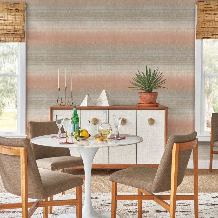 The dining room radiates rustic elegance with a round white table and four wooden chairs. A sideboard adorned with a potted plant and candles enhances the atmosphere. On the table, a green bottle, two glasses, and a fruit bowl create an inviting arrangement against the backdrop of York Wallcoverings' Desert Textile Wallpaper in Pink, which harmonizes beautifully with woven blinds.