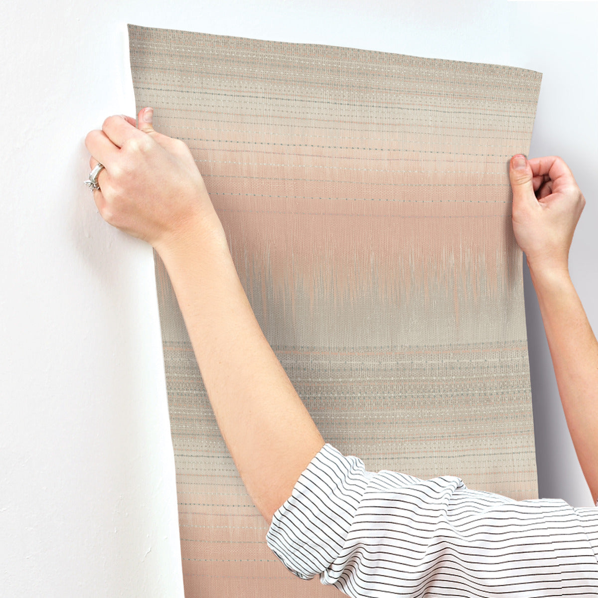 A person is hanging a piece of Desert Textile Wallpaper Pink by York Wallcoverings on a white wall. They are wearing a striped shirt, and both hands are visible, smoothing out the paper. The wallpaper showcases a subtle, earthy tone design that infuses the space with rustic elegance.