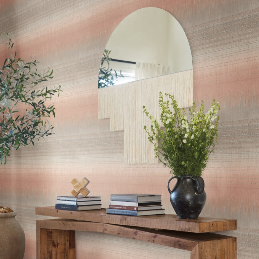 A wooden console table exudes rustic elegance, displaying a black vase with green and white flowers. Stacked books and a decorative object enhance the charm, while a large, uniquely shaped mirror on the wall above complements the scene against the Desert Textile Wallpaper in Pink by York Wallcoverings. Nearby, a potted plant breathes life into the setting.