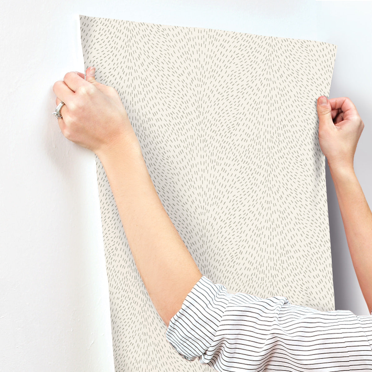 Wearing a striped shirt, a person smooths York Wallcoverings' Dew Drop Beige Wallpaper (60 SqFt), featuring small scattered lines, onto a white wall. Only their arms and hands are visible as they apply the non-woven wallpaper.