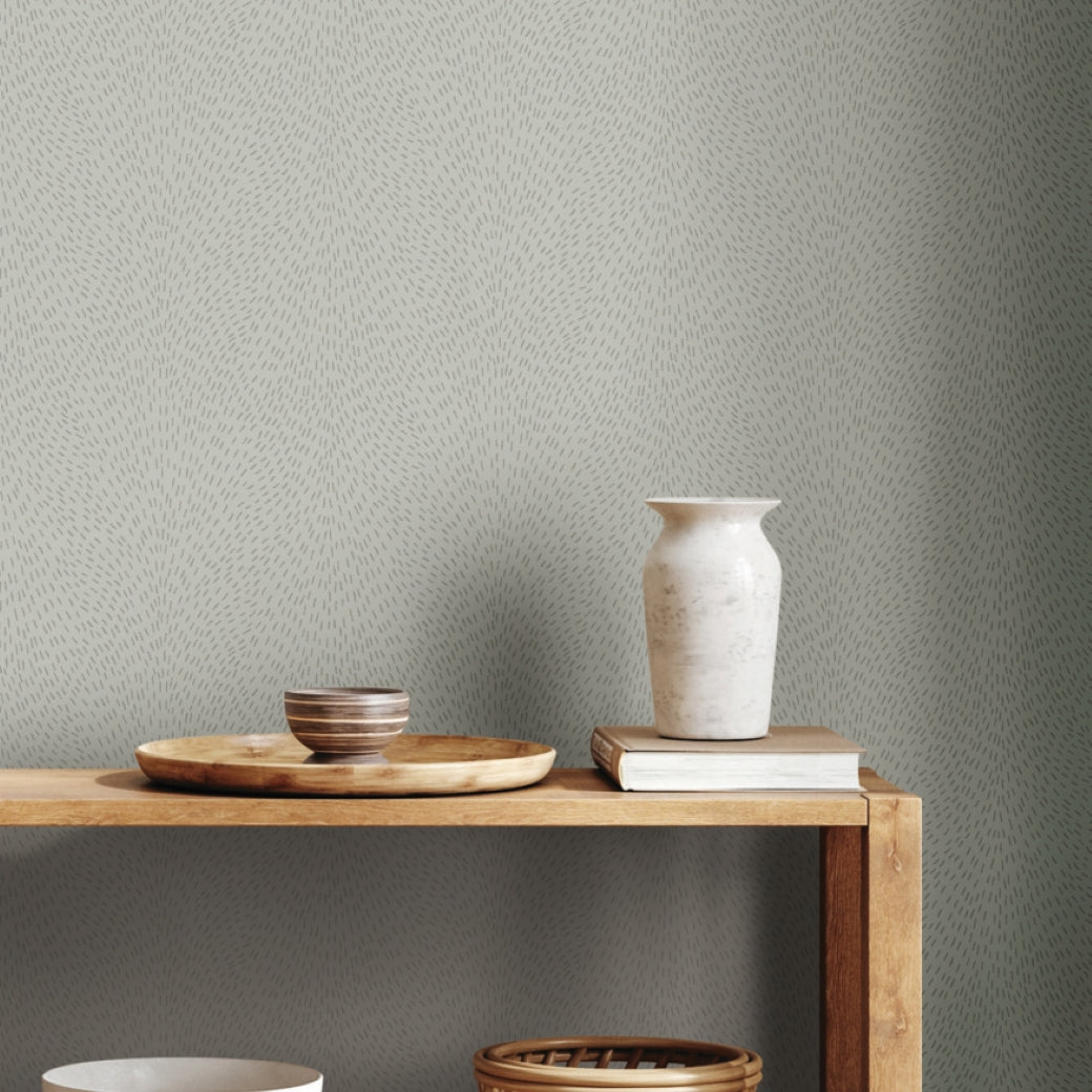 A wooden table holds a beige ceramic vase, stacked book, round tray, and two small bowls against Dew Drop Grey Wallpaper by York Wallcoverings. The scene embodies simple, minimalist style.