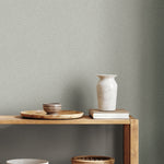 A wooden table holds a beige ceramic vase, stacked book, round tray, and two small bowls against Dew Drop Grey Wallpaper by York Wallcoverings. The scene embodies simple, minimalist style.