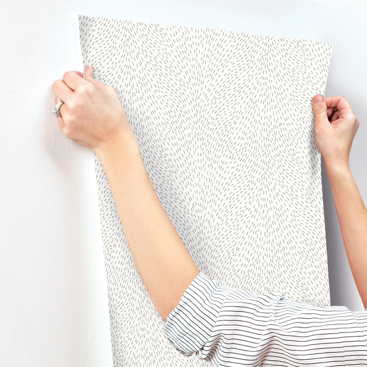 A person with rolled-up sleeves is applying York Wallcoverings Dew Drop White Wallpaper (60 SqFt), featuring thin, short lines, to a white wall, using both hands to smooth it out.