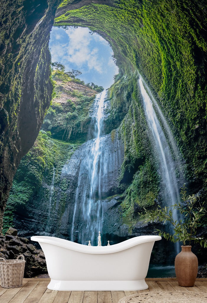 A white freestanding bathtub is positioned in front of a large Decor2Go Wallpaper Mural flowing down a lush, moss-covered rock face, viewed from a cave-like perspective with a glimpse of the sky above.