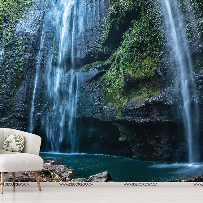 A serene indoor setting featuring a large, vivid wall mural of an Elevated Waterfall Wallpaper Mural from Decor2Go Wallpaper Mural and blue pool. An elegant chair and side table with a blanket and cushions are in the foreground, blending indoor comfort with outdoor.