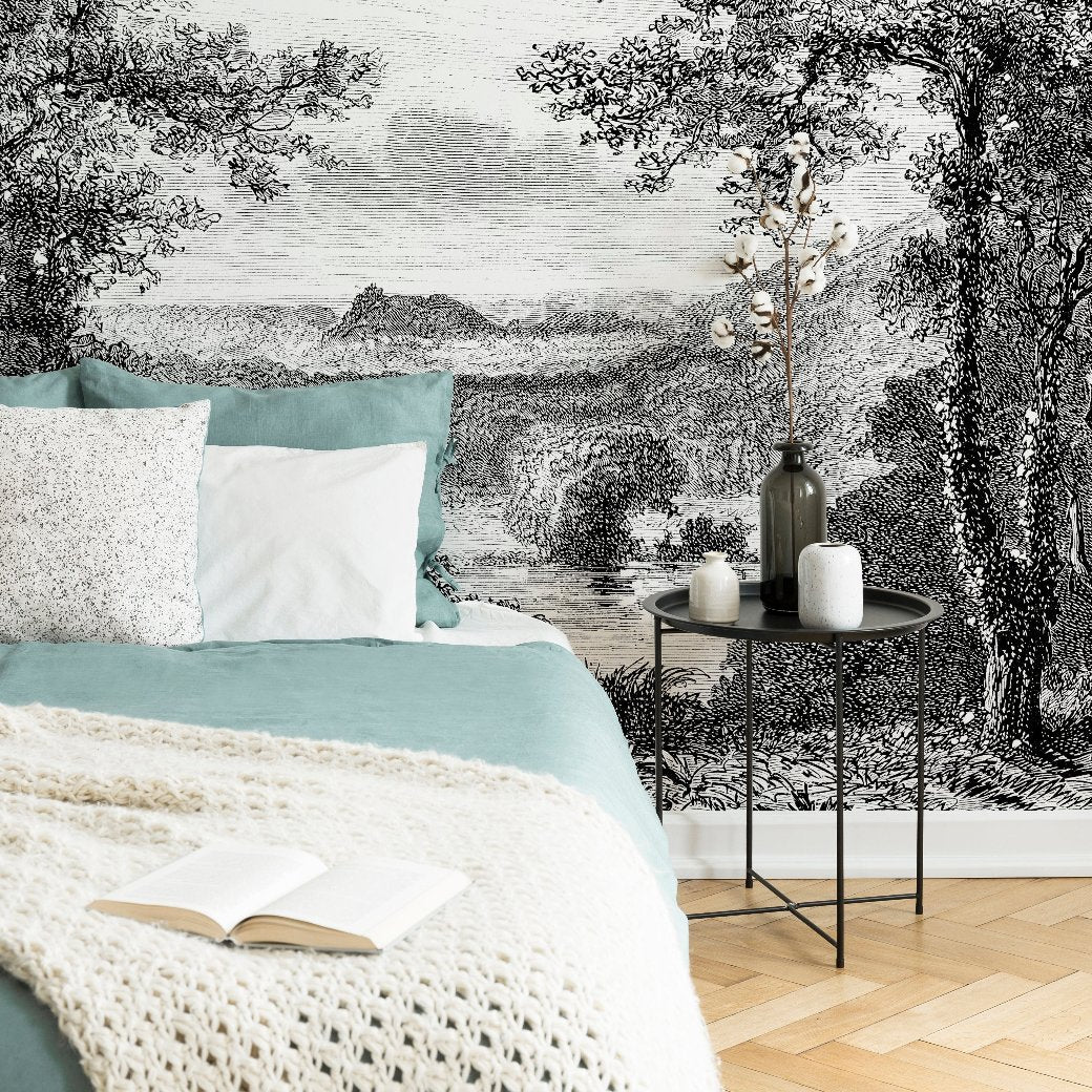 A cozy bedroom with a green and white bed and a textured blanket. A round black side table holds a vase with branches and a book. The backdrop features the Decor2Go Wallpaper Mural's Enchanted Grove Wallpaper Mural, showcasing a serene landscape of trees and mountains that exudes natural tranquility.