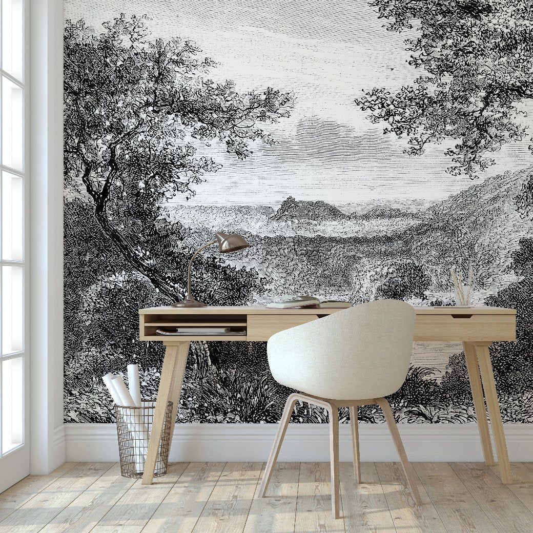 A minimalist workspace featuring a wooden desk and a white chair is complemented by the charm of the Decor2Go Enchanted Grove Wallpaper Mural. A lamp and books adorn the desk, set against this mural that showcases a lush, hilly landscape with trees and distant mountains, creating an ambiance of natural tranquility.