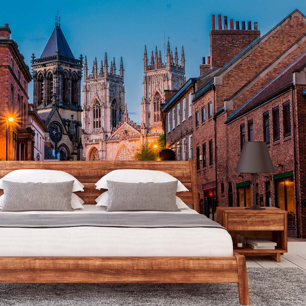 A surreal composite image of a bedroom with a large bed and wooden furniture set outdoors in an English city street, blending urban and historical architecture, including cathedrals, under a twilight sky featuring the Decor2Go Wallpaper Mural's England Stroll Wallpaper Mural.
