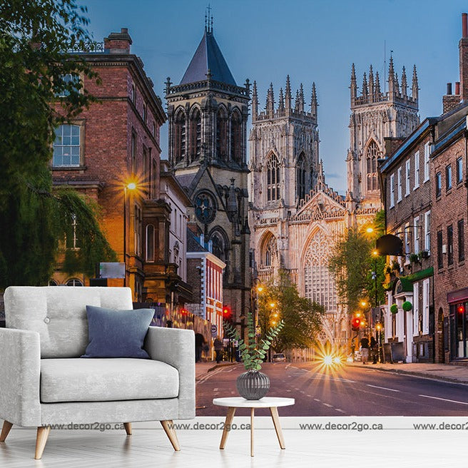 A composite image blending a city street in England at dusk, featuring lit street lamps and a historic cathedral, with a modern living room setup including a grey sofa and a small round table, all showcased on the Decor2Go Wallpaper Mural - England Stroll.