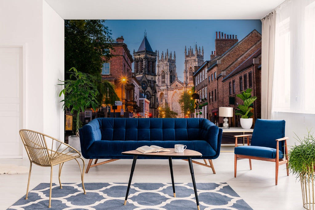 A modern living room featuring a large mural of a vibrant Decor2Go Wallpaper Mural, depicting scenic sites and historical architecture in England. The room includes a navy blue couch, blue armchair, gold accent chair, and a small.