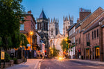 A picturesque evening view of a historic street leading to a large gothic cathedral, illuminated by street lights and visible car's light trails, framed by old buildings and a clear blue sky. This scene captures Decor2Go Wallpaper Mural's England Stroll Wallpaper Mural.