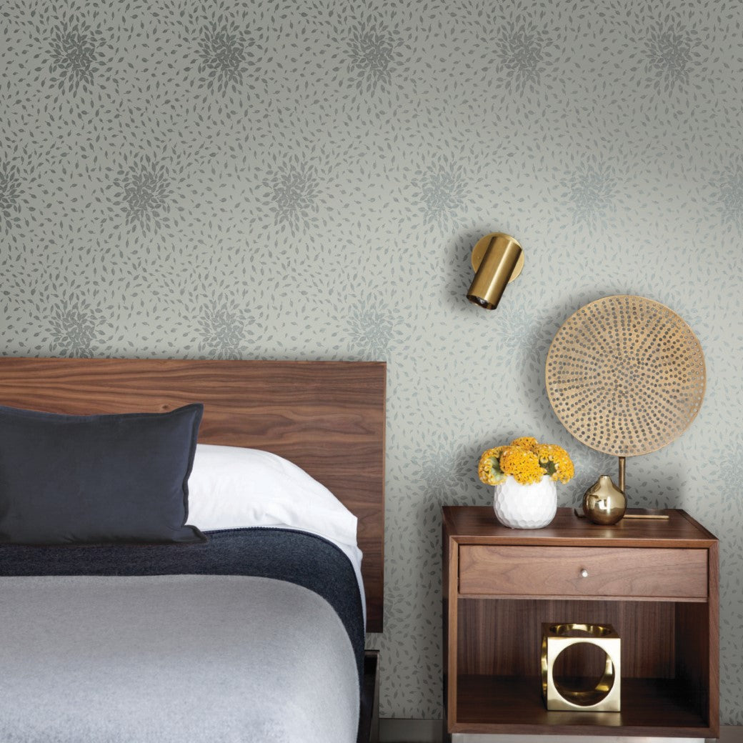 A modern bedroom showcases a wooden bed with gray bedding and a dark pillow. A wooden nightstand holds a lamp, round woven decor, and a vase with yellow flowers. Walls feature York Wallcoverings' Eucalyptus & Silver Petite Leaves Wallpaper, complemented by gold sconce from the Modern Metals Collection.