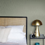 A cozy bedroom showcases a rattan headboard, white and beige pillows, and a neatly made bed. A golden dome lamp from the Modern Metals Collection sits on a black bedside table with a vase of eucalyptus and silver sprigs plus a blue candle. The wall features York Wallcoverings' Eucalyptus & Silver Sprigs Wallpaper.