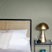 Bedroom - A cozy showcases a rattan headboard, white and beige pillows, and a neatly made bed. A golden dome lamp from the Modern Metals Collection sits on a black bedside table with a vase of eucalyptus and silver sprigs plus a blue candle. The wall features York Wallcoverings' Eucalyptus & Silver Sprigs Wallpaper