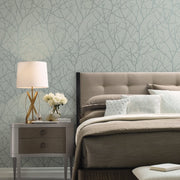 Bedroom - A serene boasts a cozy beige bed with pillows next to a nightstand adorned with a lamp, white flowers, and books. The walls feature York Wallcoverings' Eucalyptus & Silver Trees Silhouette Wallpaper, enhancing the room's cozy elegance