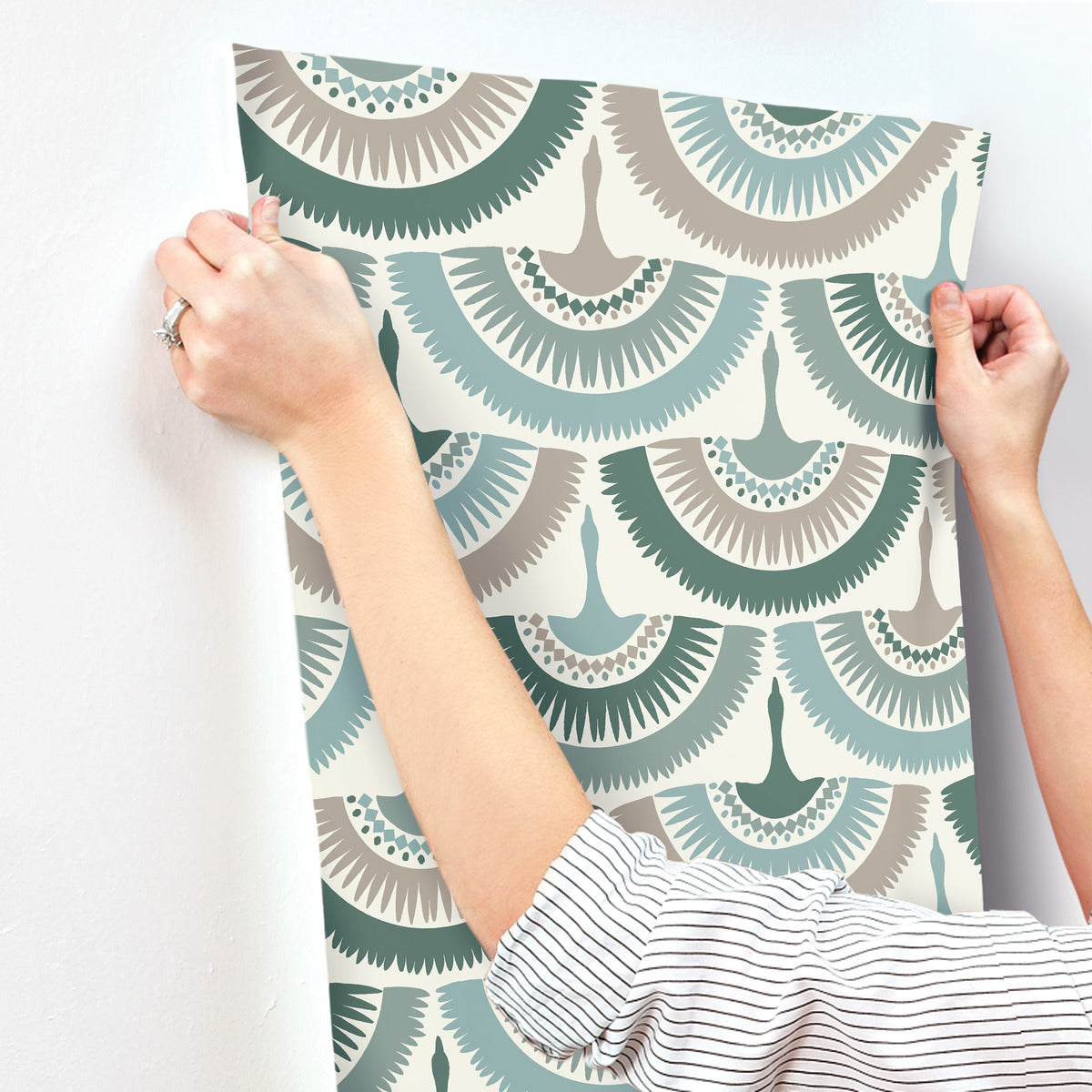 A person in a striped shirt, adorned with a ring, holds up a sheet of York Wallcoverings' Feather and Fringe Wallpaper Green against a white wall. The durable material showcases an abstract fan design in teal, beige, and gray inspired by nature.