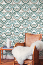 Against a wall adorned with York Wallcoverings' Feather and Fringe Wallpaper in Green, featuring a teal and cream fan pattern, sits a wicker chair draped with a white faux fur throw. Beside the chair, a small table holds a decorative white vase, complemented by the nature-inspired design of the wooden floor.