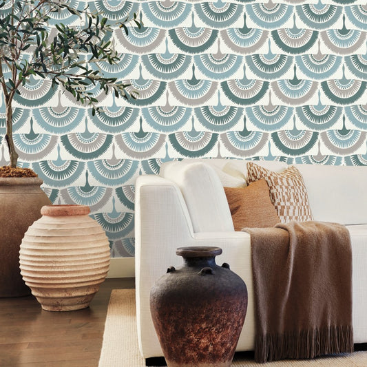 A cozy living room featuring a white sofa adorned with a brown throw and patterned cushions. An olive tree stands beside large decorative clay pots, while the York Wallcoverings Feather and Fringe Wallpaper in Green adds a teal and white Art Deco fan pattern. The wooden floor beautifully complements this nature-inspired design.