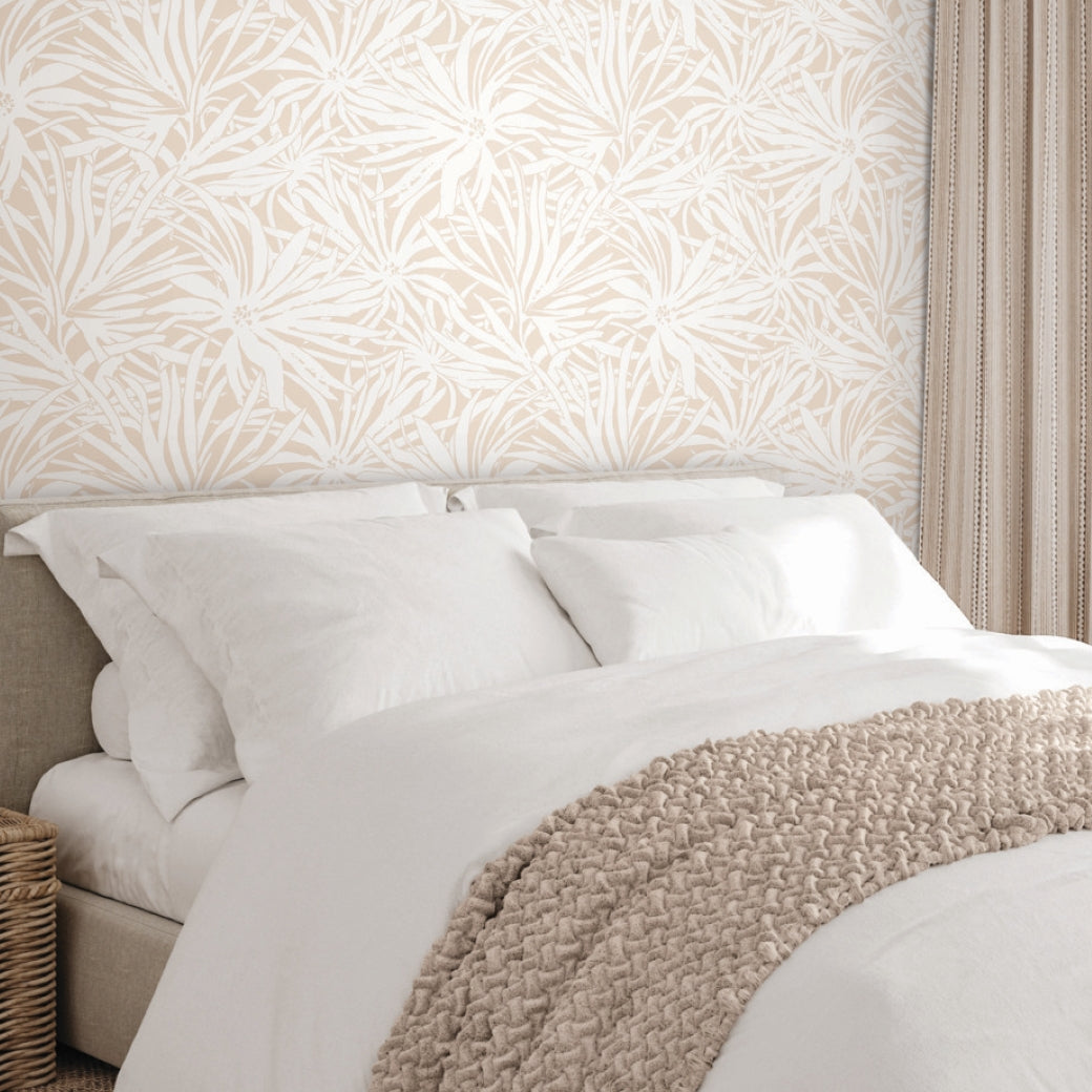 A cozy bed with white bedding and a beige knitted throw, set against York Wallcoverings' Floral Canopy Blush Wallpaper. Beige striped curtains hang nearby, and a wicker basket sits next to the bed.