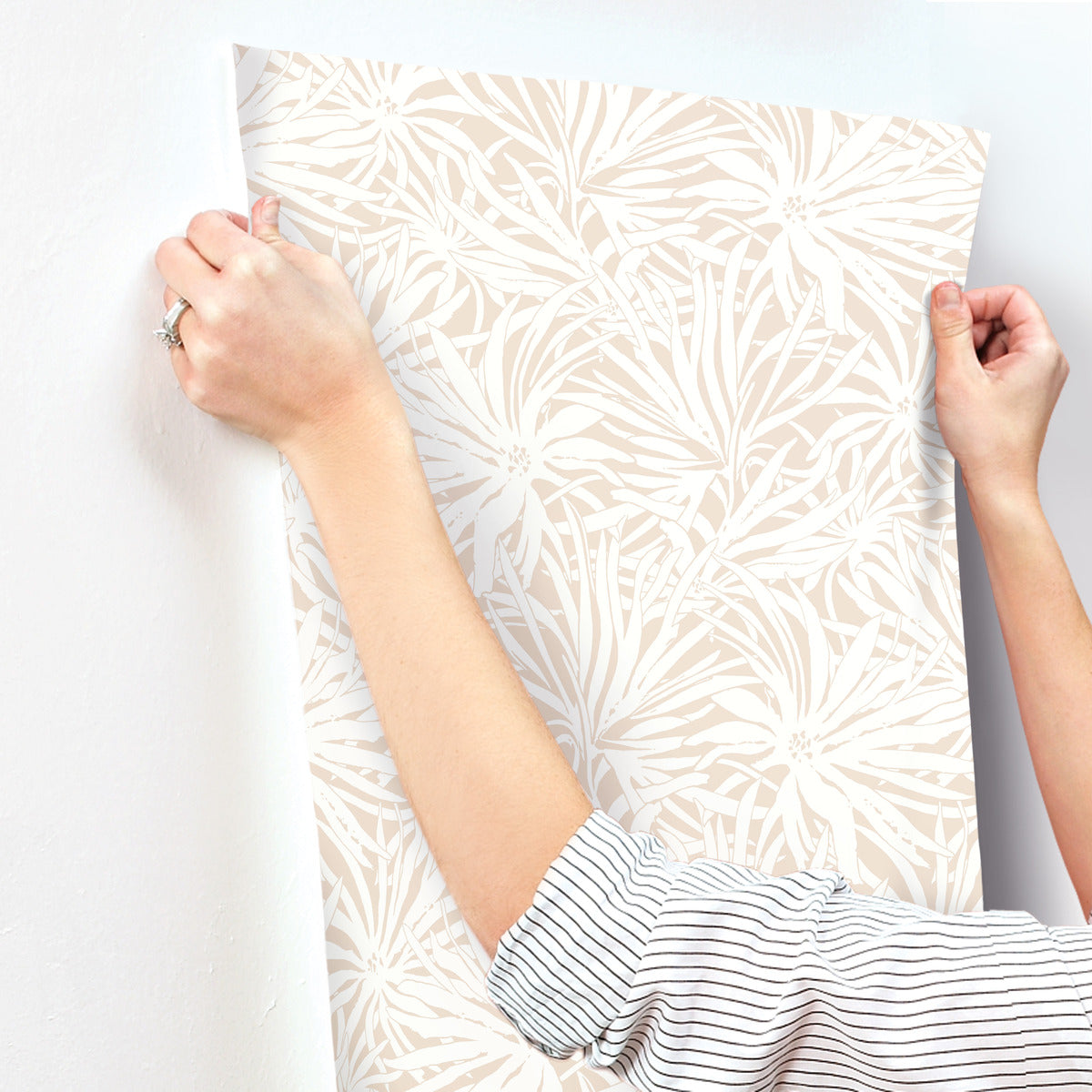 Wearing a striped shirt, someone is smoothing York Wallcoverings' Floral Canopy Blush Wallpaper (60 SqFt)—a beige and white botanical wallpaper with abstract floral designs—onto a wall. Only their hands and arms are visible.