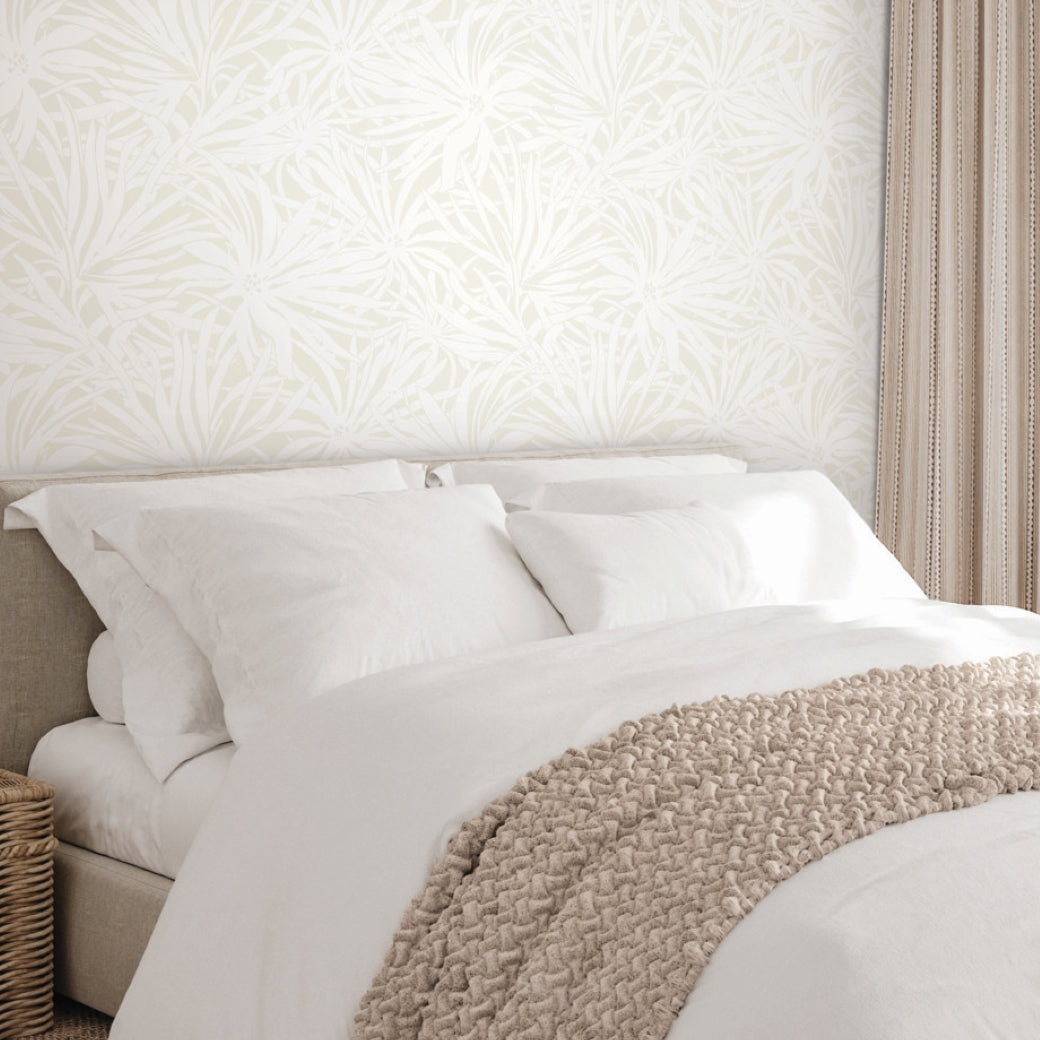 A neatly made bed with white linens and a beige throw sits against York Wallcoverings' Floral Canopy Cream Wallpaper, while beige curtains and a wicker basket nearby enhance the serene, nature-inspired look.