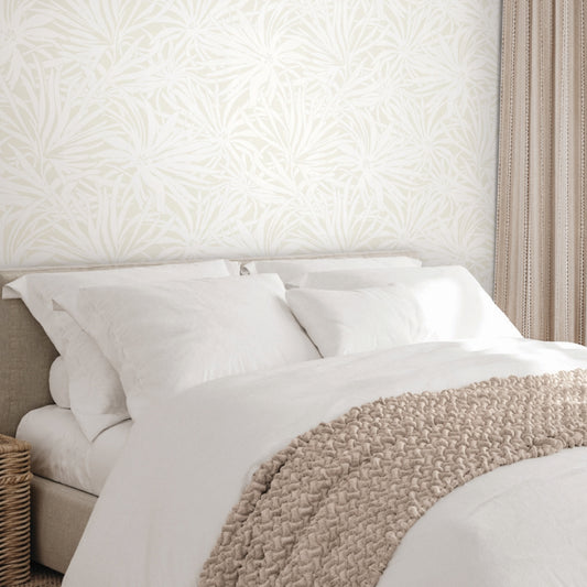 A neatly made bed with white linens and a beige throw sits against York Wallcoverings' Floral Canopy Cream Wallpaper, while beige curtains and a wicker basket nearby enhance the serene, nature-inspired look.