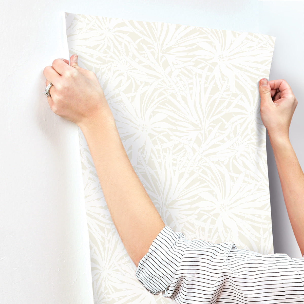 A person wearing a striped shirt is preparing to apply York Wallcoverings’ Floral Canopy Cream Wallpaper (60 SqFt), featuring a subtle leaf pattern, onto a white wall.