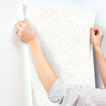 A person wearing a striped shirt is preparing to apply York Wallcoverings’ Floral Canopy Cream Wallpaper (60 SqFt), featuring a subtle leaf pattern, onto a white wall.