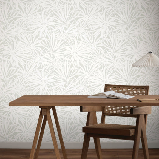 A wooden desk with an open book and lamp beside a wooden chair, set against a light wall adorned with York Wallcoverings’ Floral Canopy Grey Wallpaper (60 SqFt), featuring a subtle leafy botanical pattern.