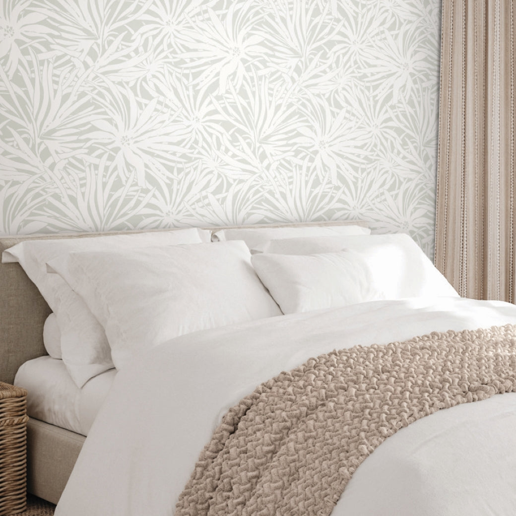A neatly made bed with white bedding and pillows sits against a wall covered in York Wallcoverings’ Floral Canopy Grey Wallpaper. A textured beige blanket is draped at the foot, and beige curtains frame the scene on the right.