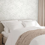 A neatly made bed with white bedding and pillows sits against a wall covered in York Wallcoverings’ Floral Canopy Grey Wallpaper. A textured beige blanket is draped at the foot, and beige curtains frame the scene on the right.