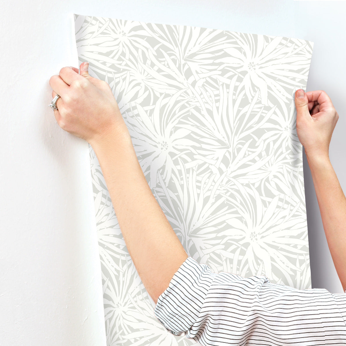 A person’s hands and forearms are shown applying York Wallcoverings' Floral Canopy Grey Wallpaper (60 SqFt), featuring a white and light gray floral design, to a white wall.