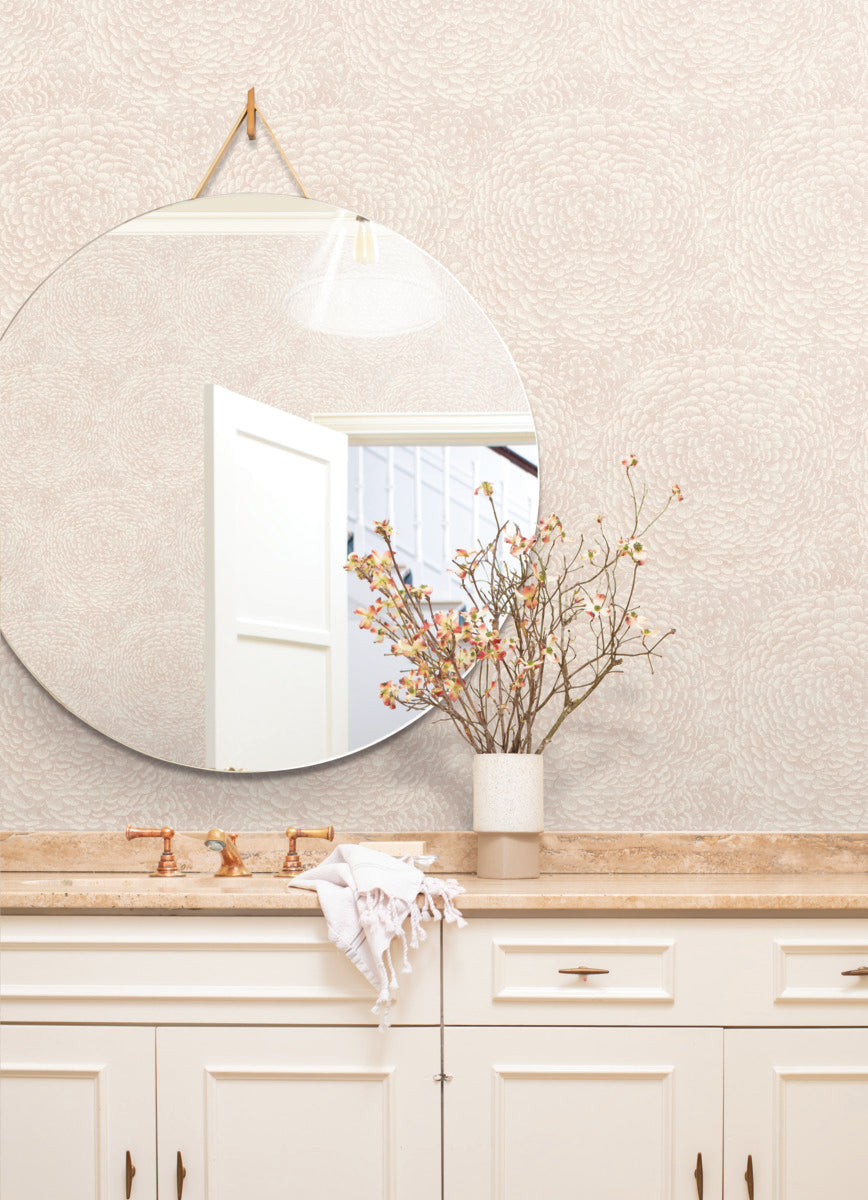 A round mirror hangs on a Floret Wallpaper Pink wall by York Wallcoverings, above a light-colored countertop, complemented by nature-inspired design elements. A white door is slightly ajar, reflected in the mirror. A vase with branches and small flowers stands on the countertop next to a folded white towel.