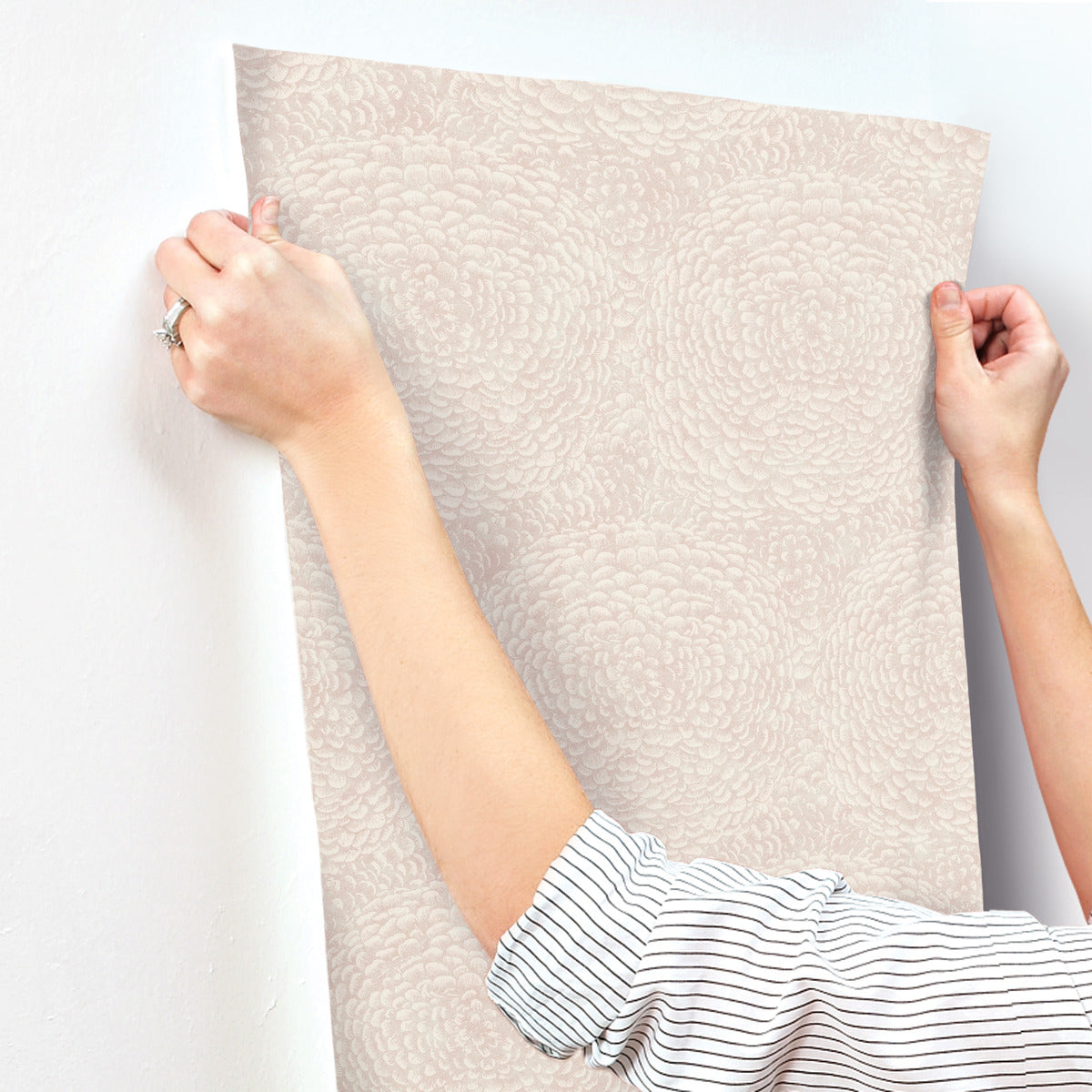 A person wearing a striped shirt applies the Floret Wallpaper Pink by York Wallcoverings to a white wall. This durable, nature-inspired design showcases subtle circular floral patterns in light beige. With gentle hands, the wallpaper is smoothed seamlessly onto the surface.
