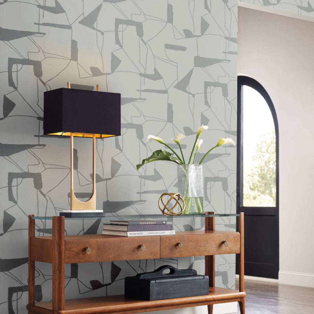 A geometric wooden console table with two drawers supports a black lamp with a wooden base, white lilies in a vase, books, and decor. The backdrop features Fog & Silver Abstract Geo Wallpaper by York Wallcoverings and is illuminated by an arched window.