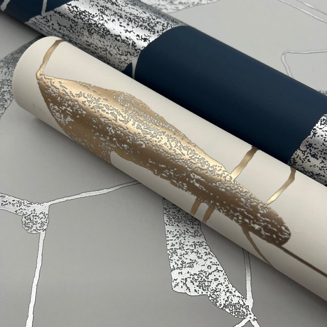 Two rolls of metallic wallpaper from the Modern Metals Collection include a dark blue and silver pattern, and a beige design with gold and silver accents, both elegantly resting on York Wallcoverings' Fog & Silver Abstract Geo Wallpaper.