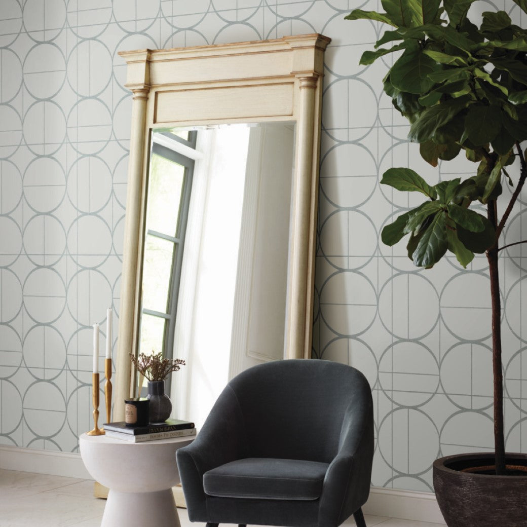 A stylish room showcases a large, framed mirror against York Wallcoverings Fog & Silver Sun Circles Wallpaper. Nearby, a dark gray armchair accompanies a small table with candles and books. Metallic accents enhance the decor, while a tall plant adds greenery.