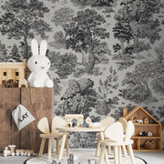 A children's playroom adorned with whimsical woodland decor, including the Forest Reverie Wallpaper Mural by Decor2Go, features wooden chairs around a table, a large white rabbit toy, and various play items like blocks and a dollhouse on wooden shelves.