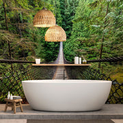 A luxurious freestanding bathtub set on a concrete floor with a backdrop of a custom Decor2Go Wallpaper Mural depicting a lush green forest and a wooden suspension bridge. Above the tub hang two woven pendant lights, enhancing