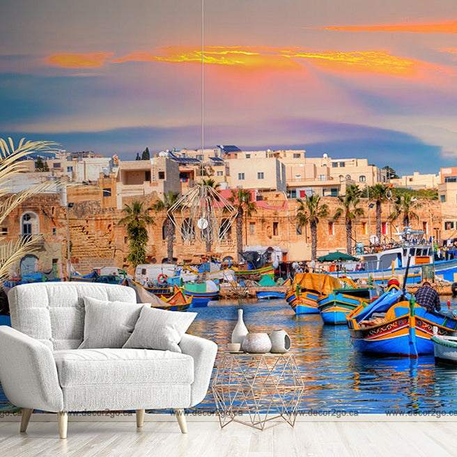 A vibrant mural of a Mediterranean coastal scene adorns a living room wall, featuring colorful boats and historic buildings under a sunset sky. The stylish white armchair and vase complement the Decor2Go Wallpaper Mural.