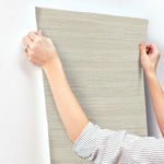 A person with a striped shirt is applying beige Fountain Grass Smokey Blue Wallpaper Blue (60 Sq.Ft.) by York Wallcoverings to a white wall. They are aligning the natural aesthetic wallpaper with their hands. The focus is on their hands holding the edges for an easy installation. A ring is visible on one of their fingers.