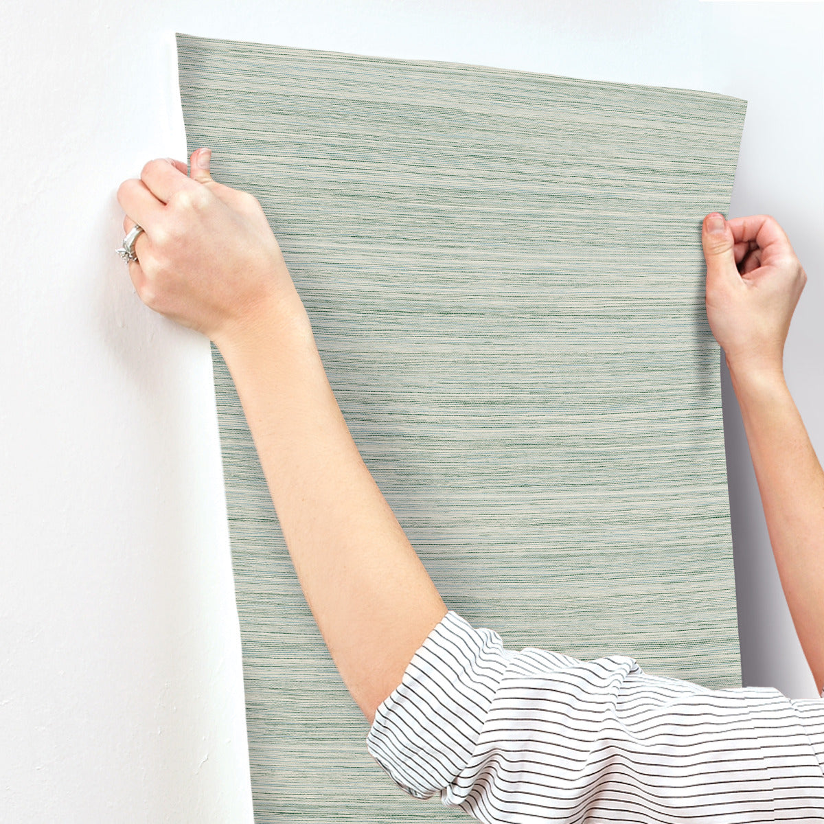 A person wearing a striped shirt is positioning a piece of York Wallcoverings Fountain Grass Camel Wallpaper Brown (60 Sq.Ft.) on a white wall. Both hands are visible, holding the wallpaper at the top corners, carefully aligning it for easy installation. A ring is visible on one of the person's fingers, emphasizing the natural aesthetic.