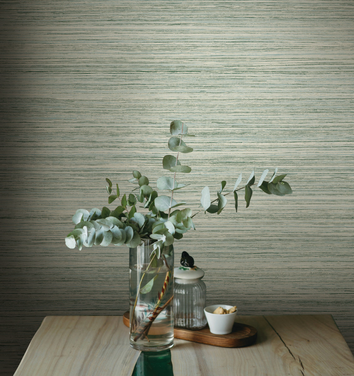 A glass vase filled with eucalyptus branches sits on a wooden table against a textured, removable York Wallcoverings Fountain Grass Smokey Blue Wallpaper Blue (60 Sq.Ft.) in neutral tones. Next to the vase is a small jar with a lid and a white bowl containing a few nuts. The scene embodies a natural aesthetic, simple and serene.